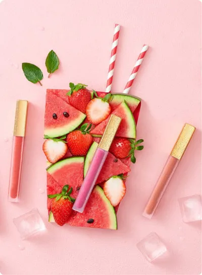 Vibrant cosmetic photography featuring lip gloss products styled with fresh watermelon and strawberries, ideal for a fun beauty brand.